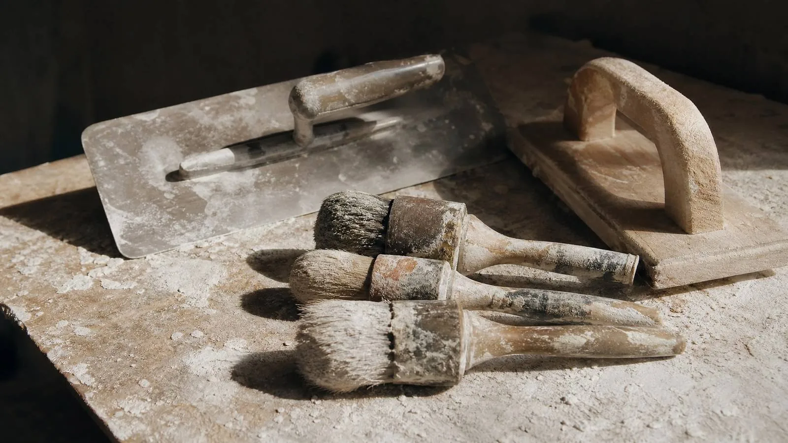 A steel plasterer's trowel, worn round brushes and a wooden float on a bench dusted with lime plaster
